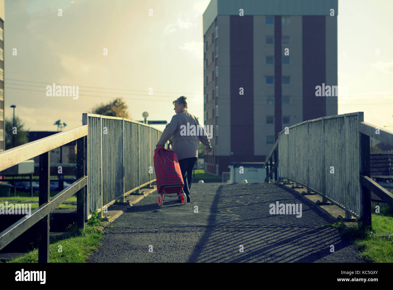 Una vecchia donna con carrello con borsa per la spesa in tartan che attraversa il ponte e il bosco di cavalieri del Clyde Canal da dietro, vista posteriore illuminata Foto Stock