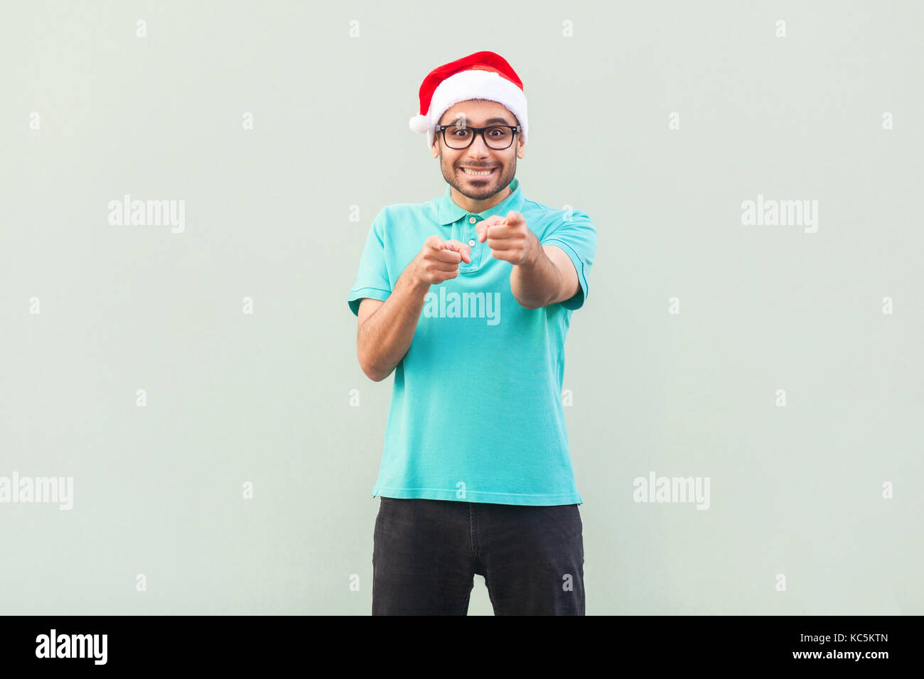 Hey! Il suo uomo di Natale! Uomo Barbuto in santa cappello e occhiali, puntare il dito e guardando la telecamera con sorriso toothy. su sfondo grigio. interna, Foto Stock