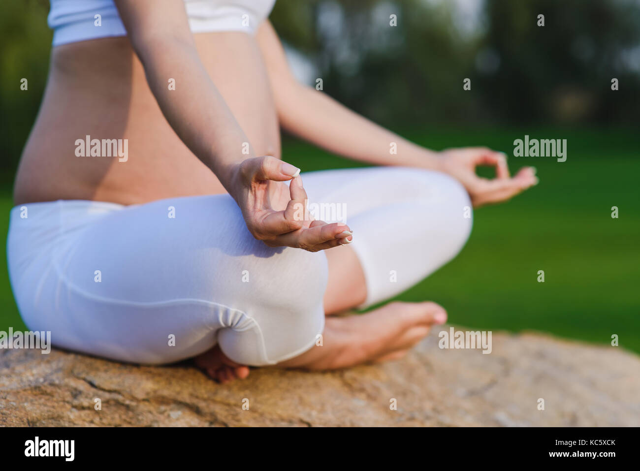Donna incinta in meditazione pongono su pietra erba nel parco. close-up di gambe incrociate di femmina con mano sul suo ginocchio. prato verde e cielo nella parte posteriore Foto Stock