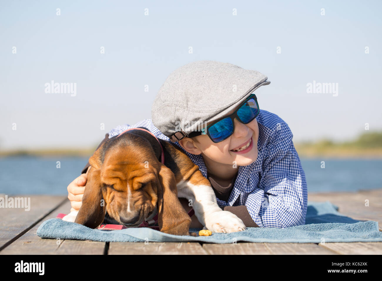 Poco carino ragazzo con un cappello e occhiali da sole giacente presso il fiume con il suo cucciolo. Essi godere insieme su una bella e soleggiata giornata. cresce, l'amore per un Foto Stock
