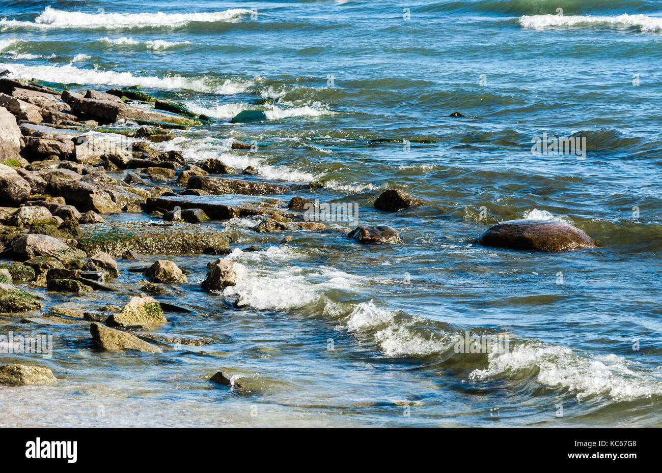 Spiaggia rocciosa con piccole onde in arrivo la rottura e la zangolatura sul bagnato pietre. Foto Stock