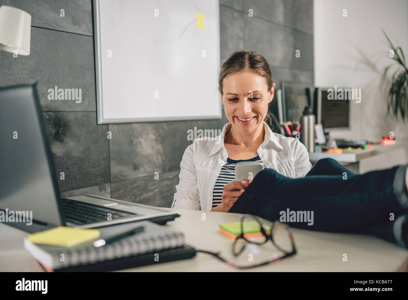 Donna che indossa una camicia bianca seduto alla scrivania in ufficio e utilizzo di smart phone Foto Stock