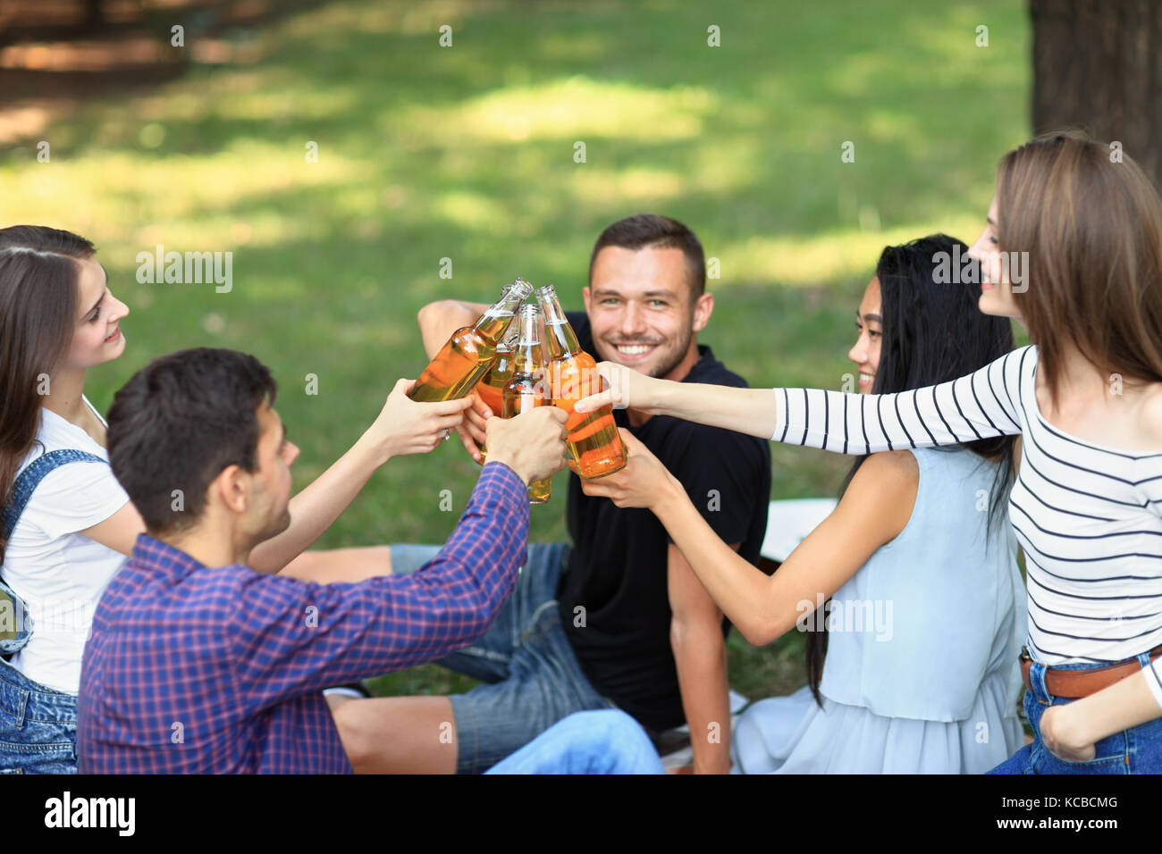 Cinque giovani felici amici clink bottiglie di birra, ridere e divertirsi in natura. allegro le ragazze ed i ragazzi in casuale vestiti estivi seduto sull'erba ho Foto Stock