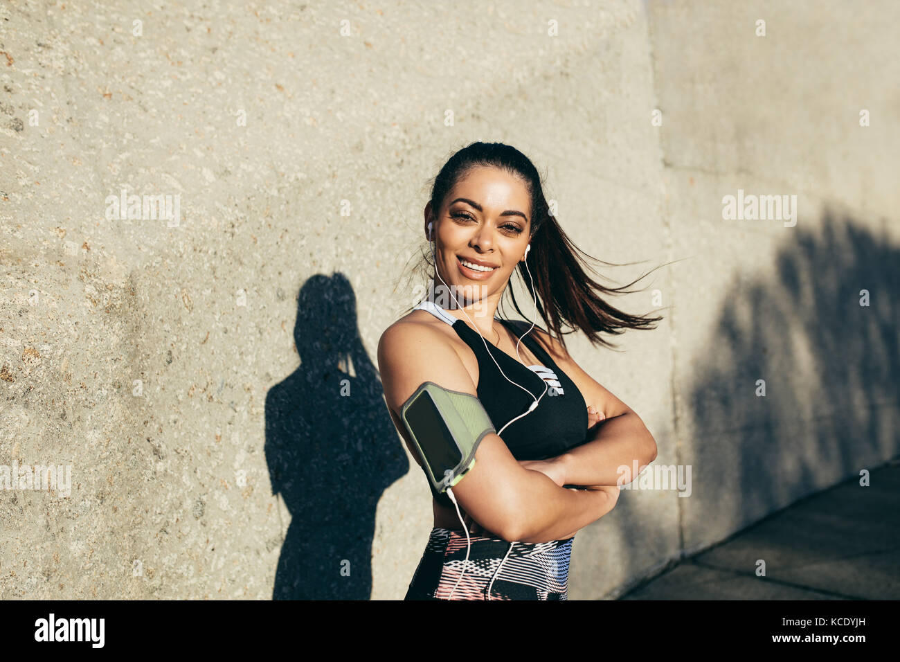 donna fitness in piedi all'aperto con le braccia incrociate e sorridente. Ritratto di una donna sportiva che indossa la fascia da braccio dello smartphone pronta per il fitness all'aperto Foto Stock