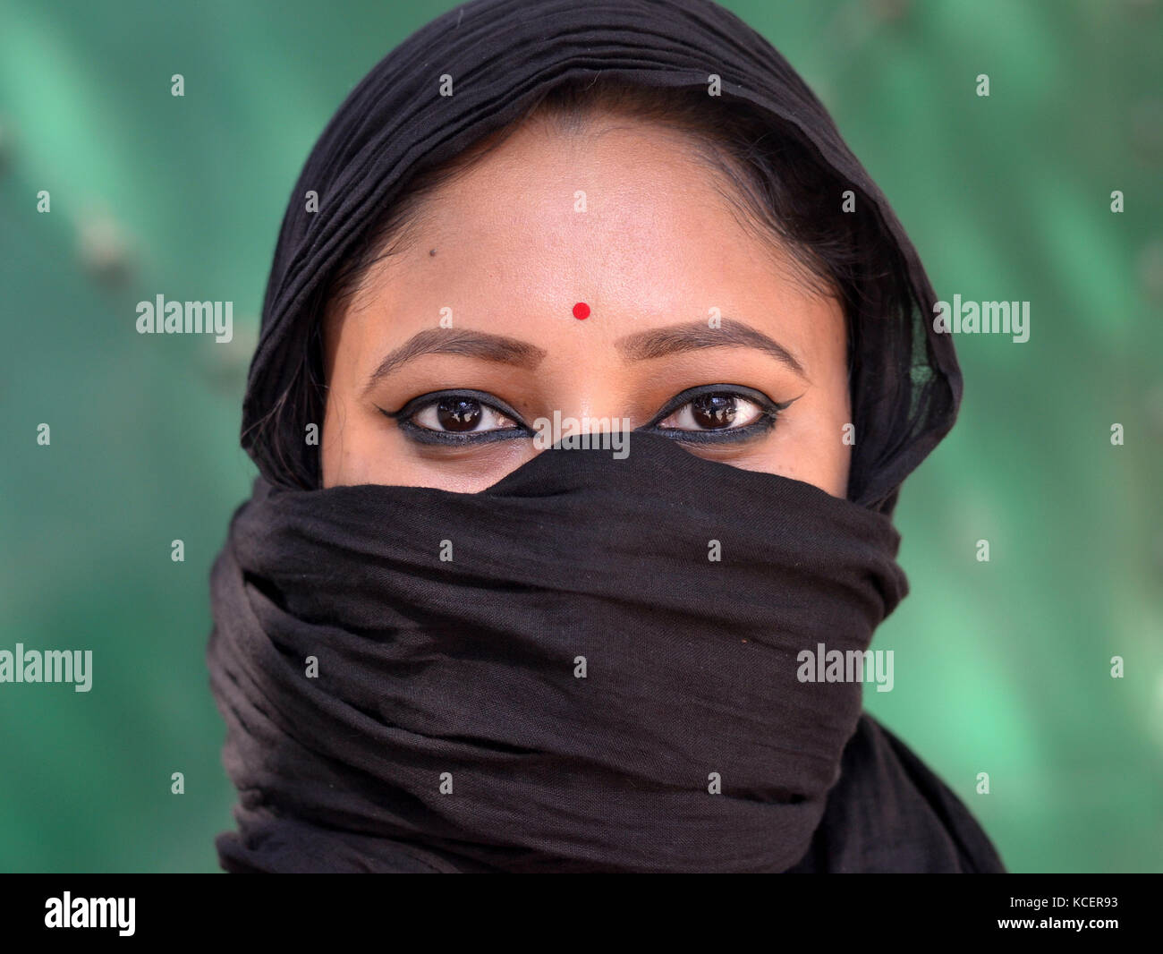 Vista a faccia intera di una giovane bellezza indù assamese con occhi a forma di mandorla, coprendo i capelli e il viso con un secolare e trendy velo nero Foto Stock