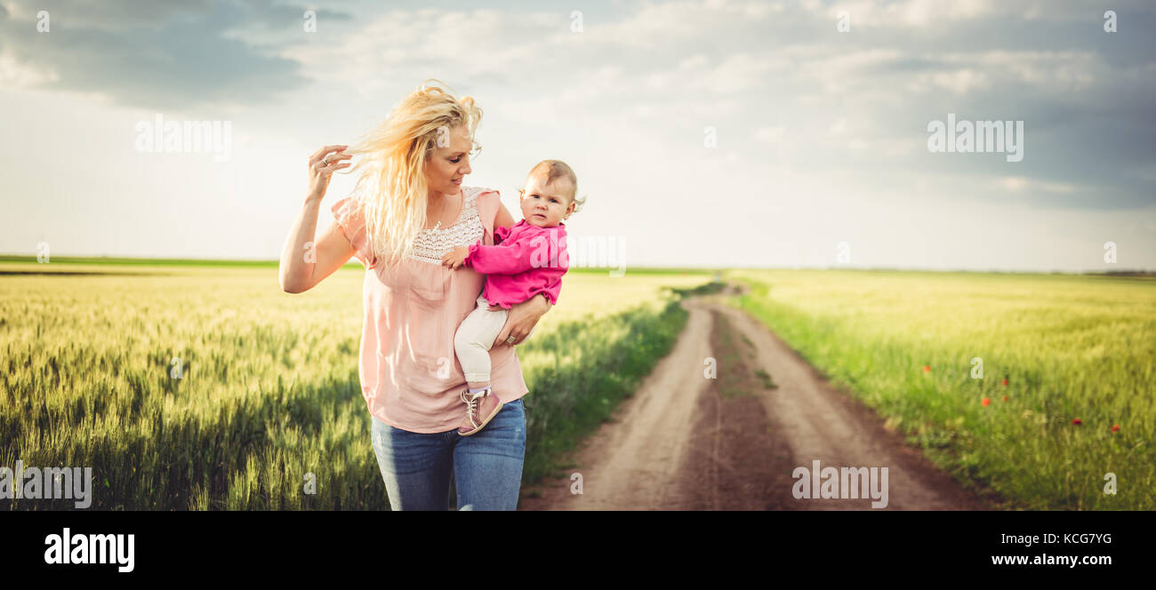 Madre tenendo la sua bambina tra le sue braccia e camminare sulla strada di ghiaia dai campi di grano Foto Stock