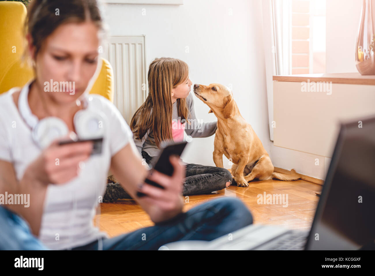 Bambina gioca con il suo cane a casa mentre la madre lo shopping online con smart phone Foto Stock