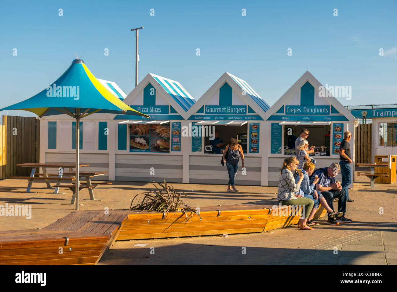 I turisti seduti a mangiare gelati e crepes di fronte alle capanne dei fast food di Hastings dai colori vivaci Foto Stock