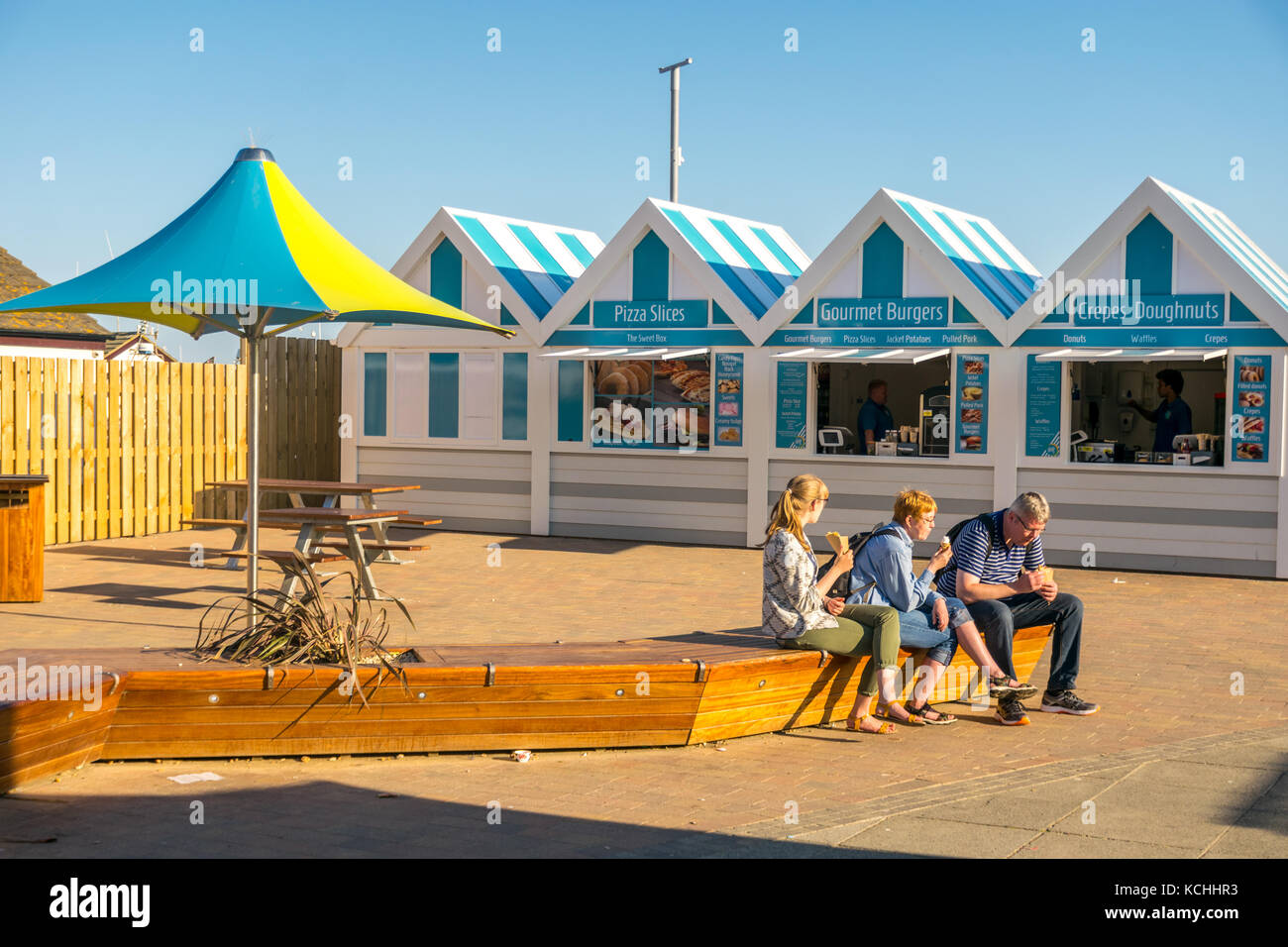 I turisti seduti a mangiare gelati e crepes di fronte alle capanne dei fast food di Hastings dai colori vivaci Foto Stock