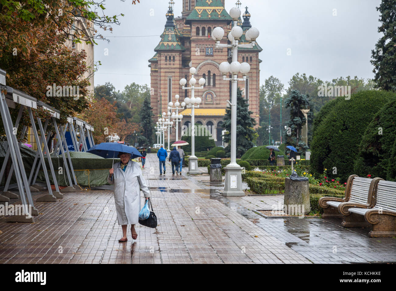 TIMISOARA, ROMANIA - 21 SETTEMBRE 2017: Una vecchia donna con un ombrello che cammina sull'iconica Piazza della Vittoria (Piata Victorei), la cattedrale metropolitana Foto Stock