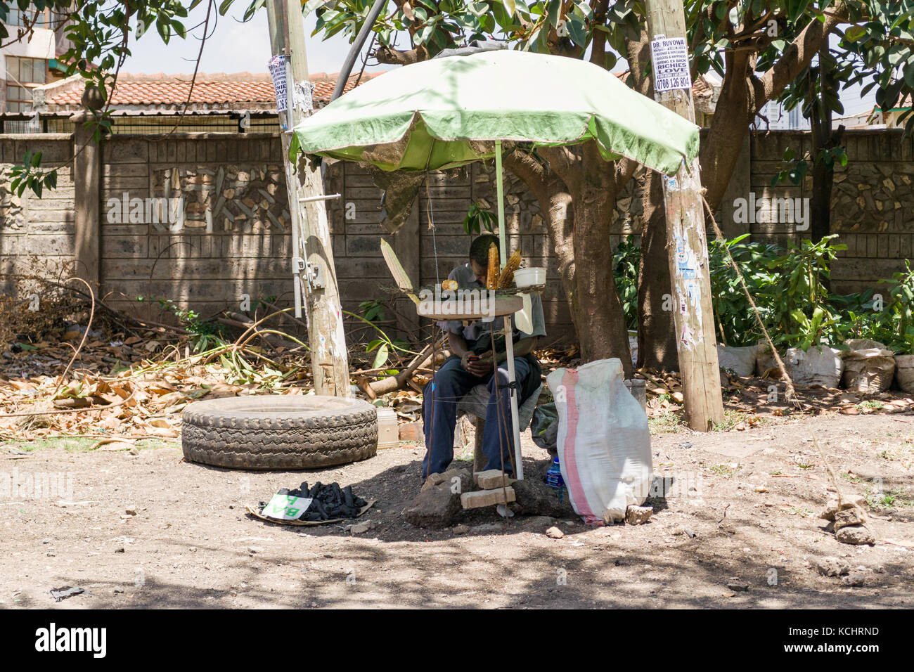 Uomo seduto in ombra sotto ombrellone vendita di mais cotta sulla pavimentazione stradale, Nairobi, Kenia Foto Stock