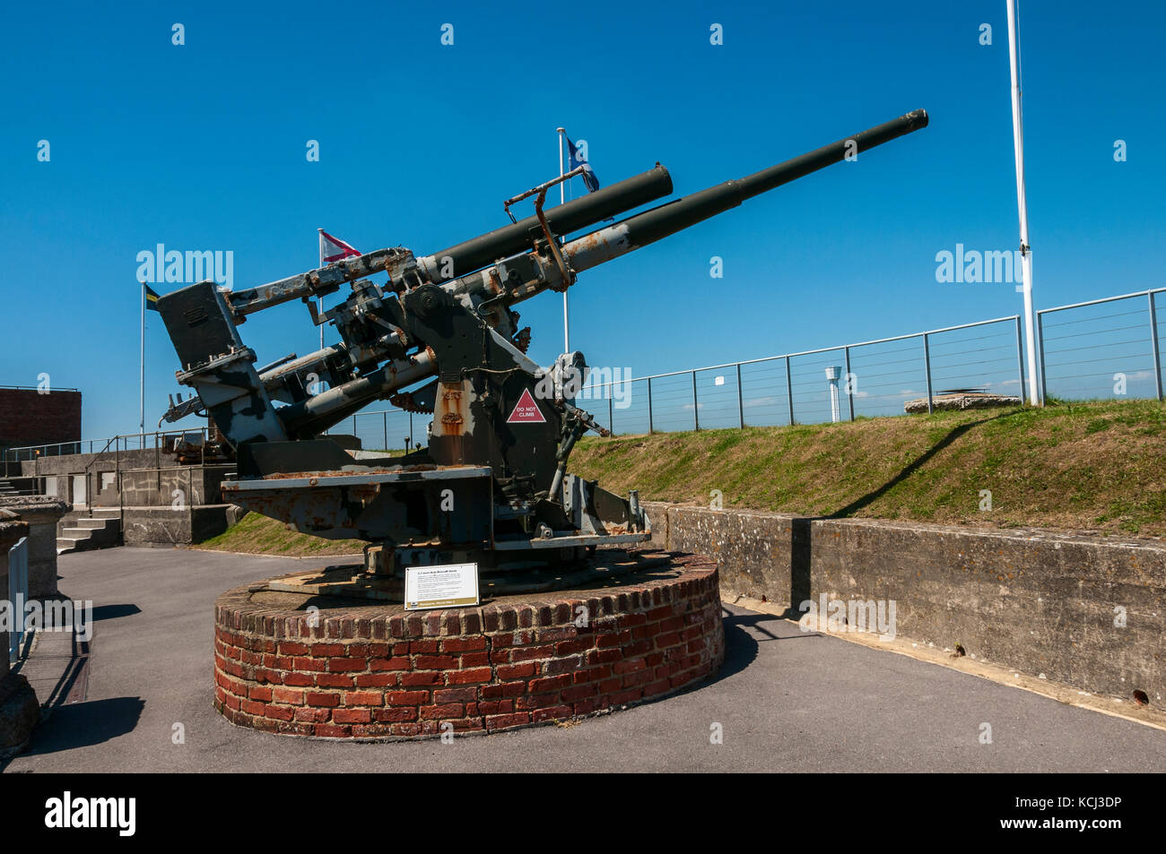 Un poco arrugginimento 3.7 pollici anti-aerei pistola appoggiata su una circolare in mattoni rossi plinto inondata di sole punti su una banca erbosa verso un cielo blu Foto Stock