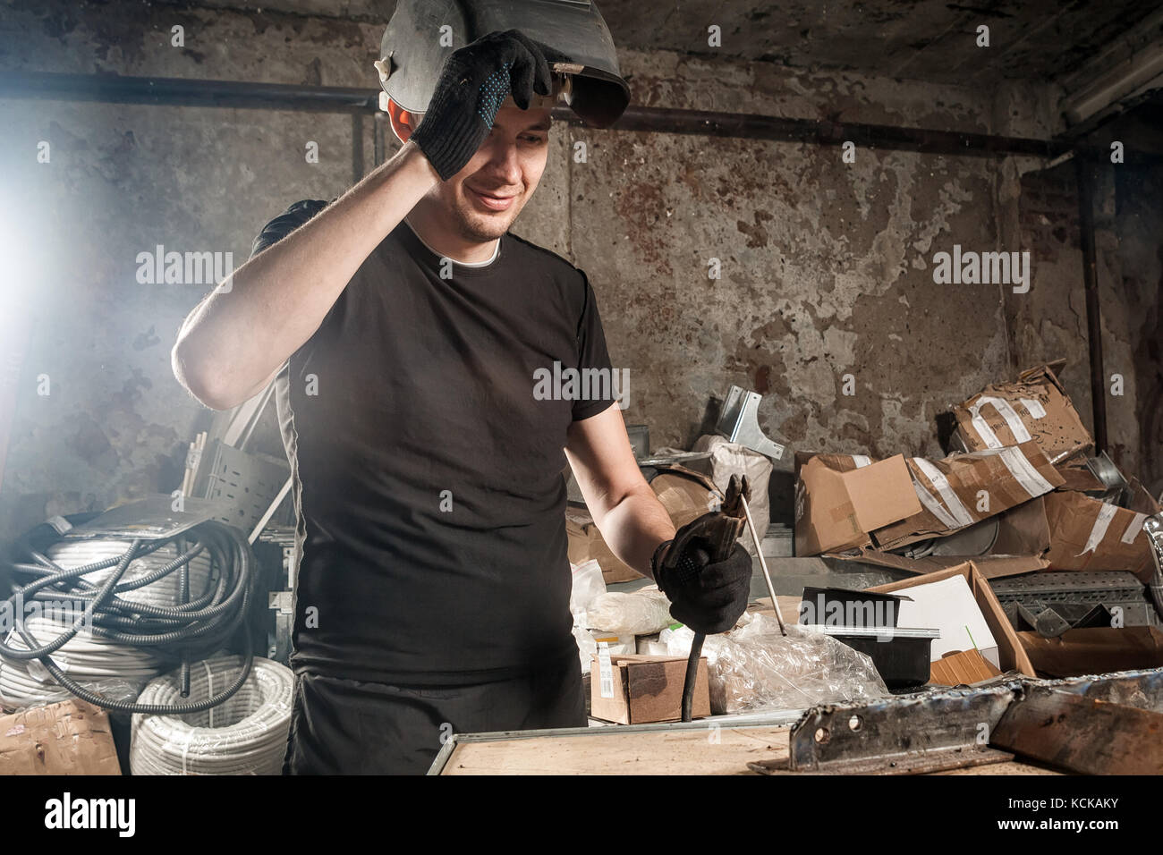 Un giovane uomo in una t-shirt nera, un nero maschera di saldatura sorrisi e prepara un metallo macchina saldatrice in una officina scuro, in background di apparecchiature, tubo flessibile Foto Stock