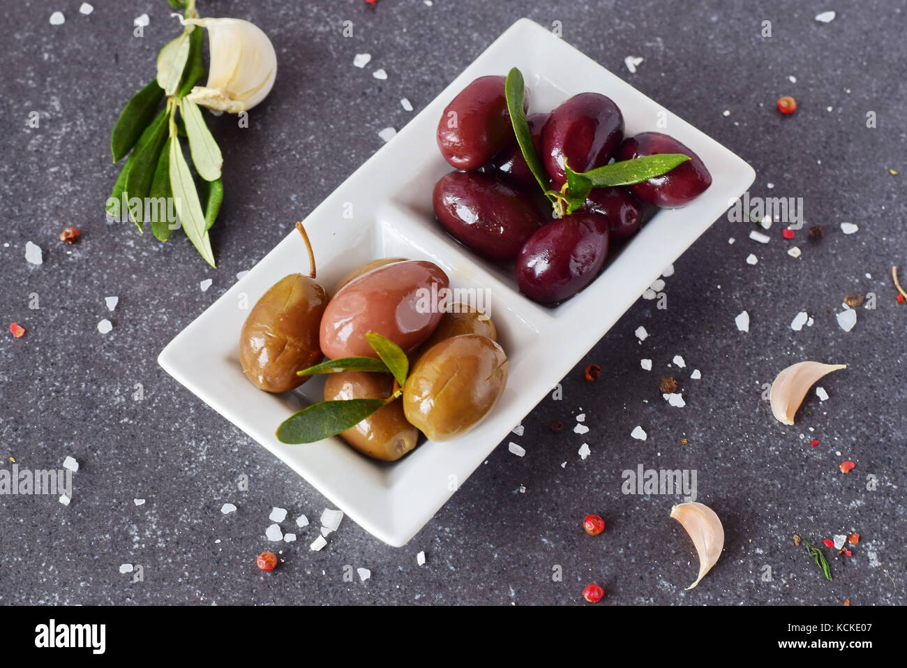 Olive nere e verdi in una piastra bianca su un grigio Sfondo astratto con limone, aglio e spezie. cucina mediterranea. Il mangiare sano concetto Foto Stock