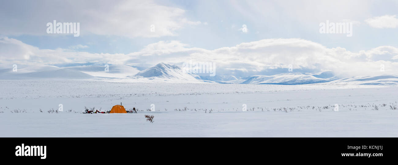 Gli snowmobilers accampano sotto le montagne McKenzie che si affacciano su un altopiano di tundra nel mezzo della stretta invernale vicino a Dechenla Lodge lungo il Canol Heritage Trail, territori del Nord Ovest, Canada Foto Stock