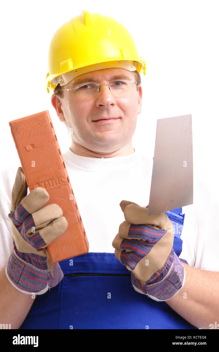 Operaio edile indossare tuta blu e giallo casco tenendo in acciaio inox cazzuola e mattone su sfondo bianco Foto Stock