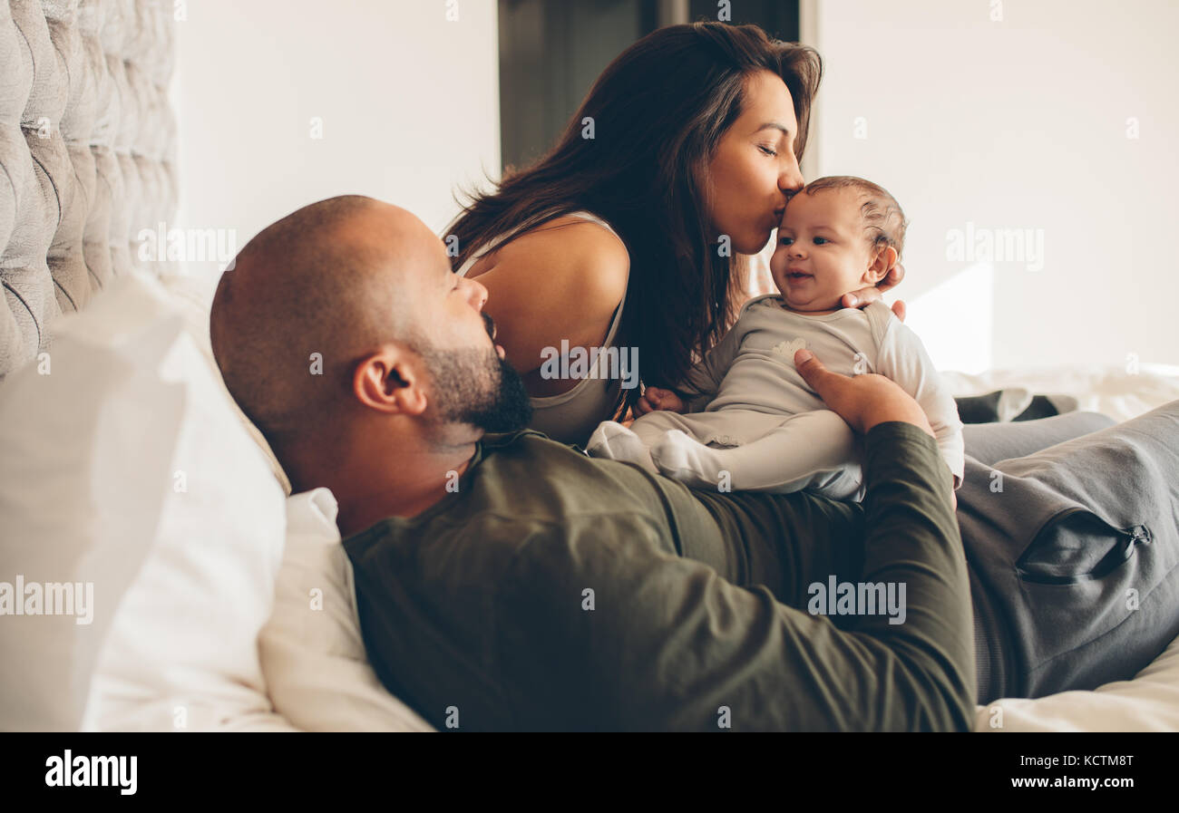 I genitori con i loro neonato ragazzo su letto di casa. Donna baciare il figlio seduto con il padre. Foto Stock