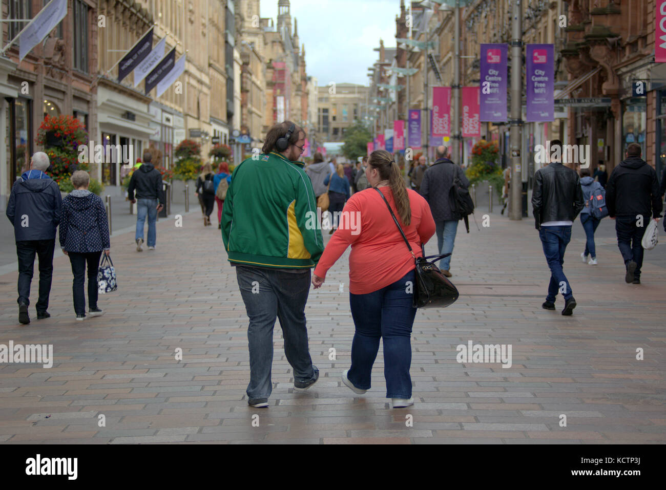 Grande coppia che ascolta musica tenendosi per mano grasso sovrappeso camminando malsana Buchanan Street style Mile Glasgow Foto Stock