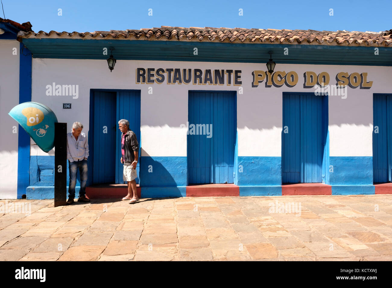 Due uomo di mezza età parlando accanto a un'Oi telefono pubblico, telefono a gettoni, chiamato orelhao, su un marciapiede di fronte ad un ristorante in Catas Altas, Minas Gera Foto Stock