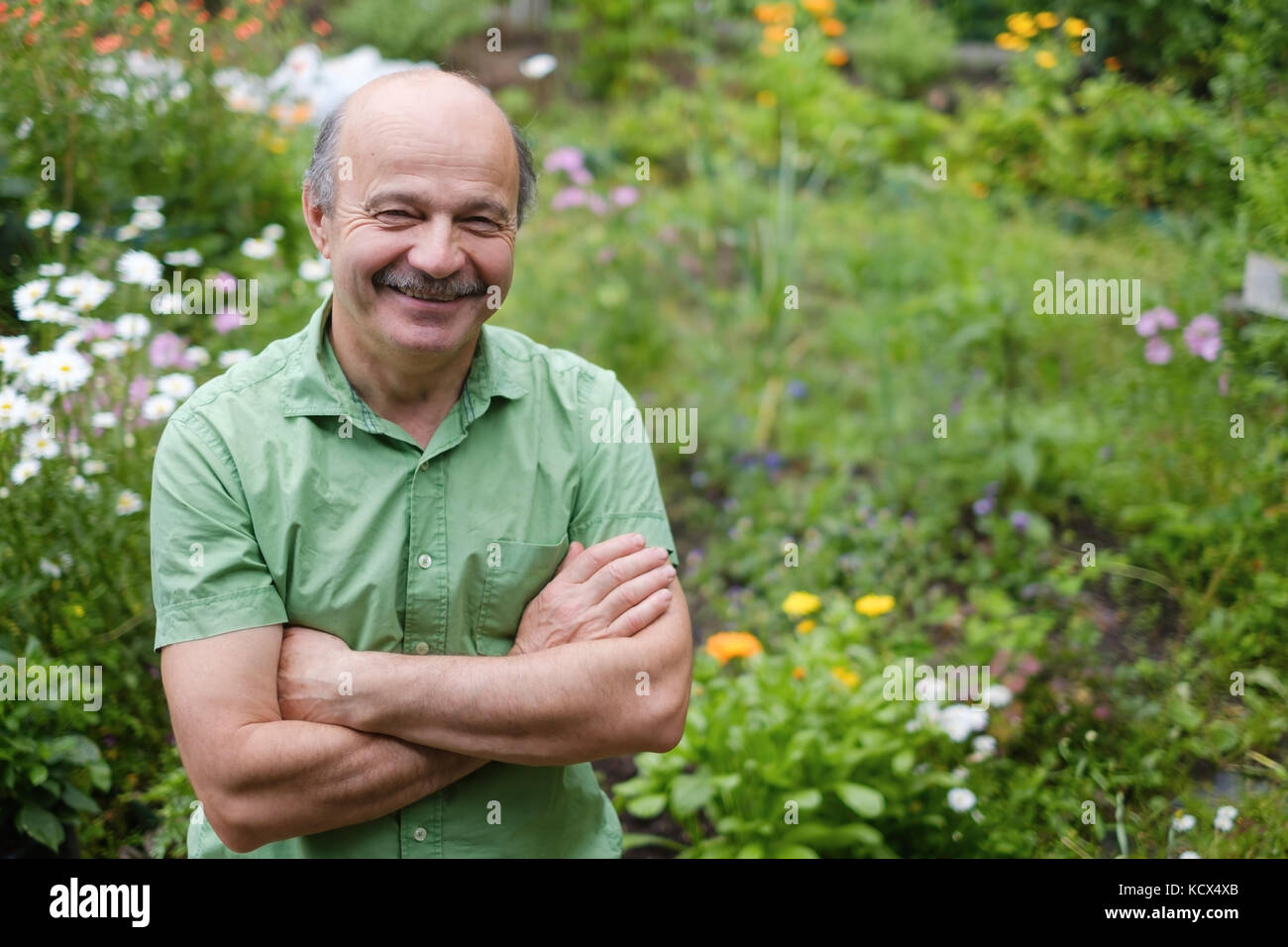 Un uomo anziano con un baffi e un bald spot in una verde t-shirt è in piedi tra i fiori nel giardino estivo, con le braccia incrociate e sorridente. Foto Stock
