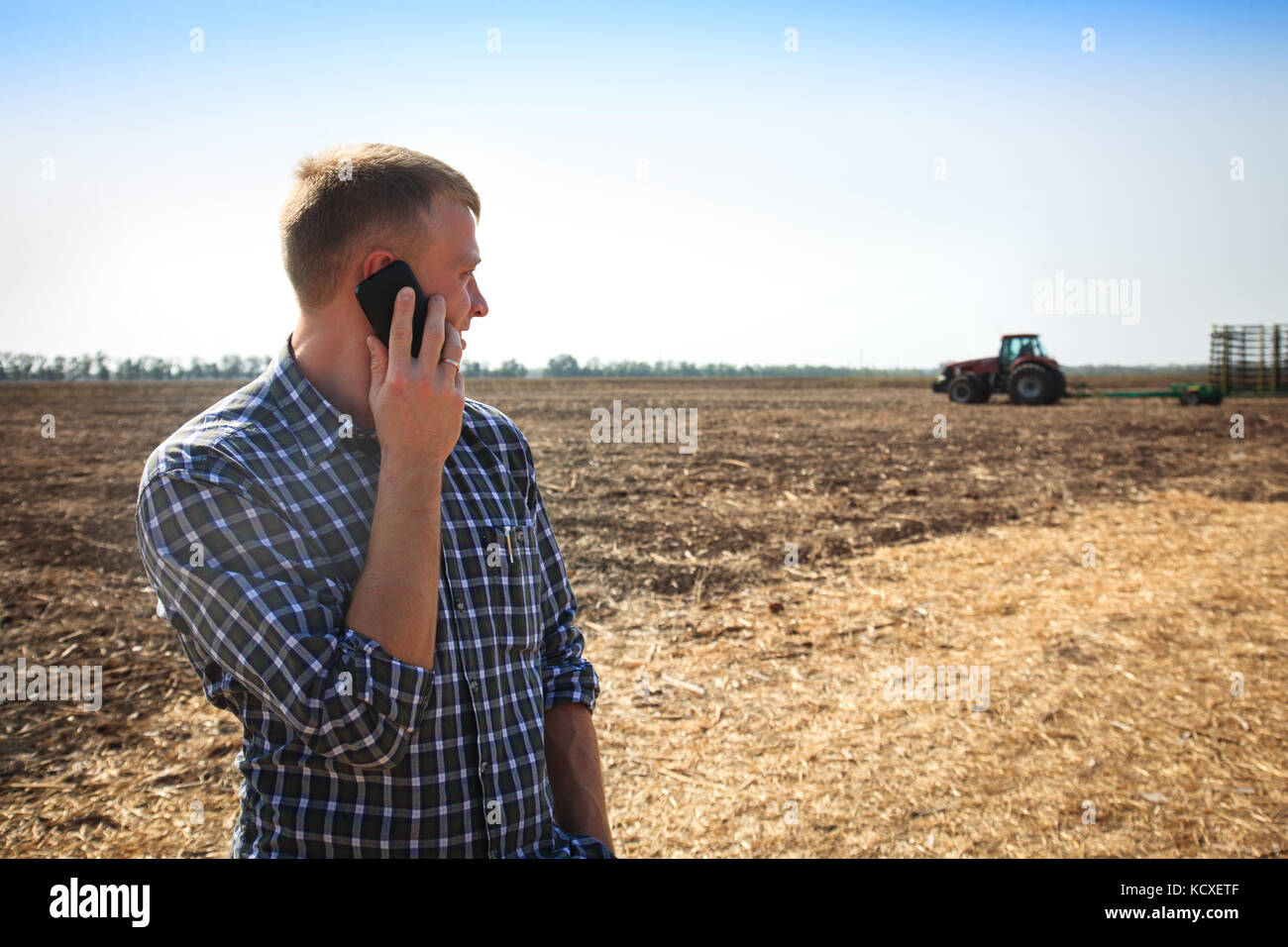 Giovane uomo con il telefono in un campo e un trattore su uno sfondo. Concetto di agricoltura e di campo funziona. Foto Stock