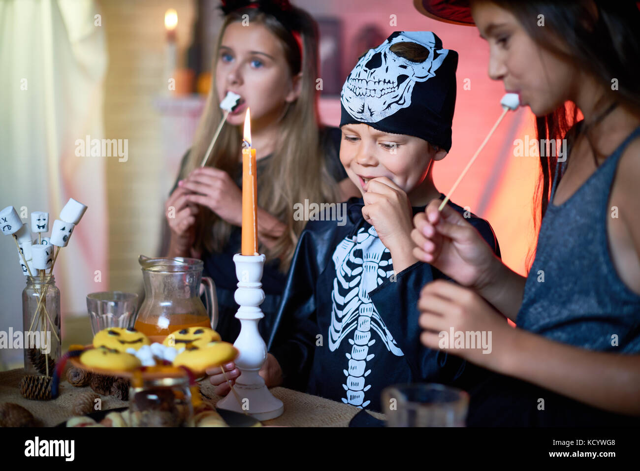 Ritratto di bello piccolo ragazzo che indossa il costume di scheletro godendo fritti gustosi marshmallows mentre celebra Halloween con amici di sesso femminile, interni o Foto Stock
