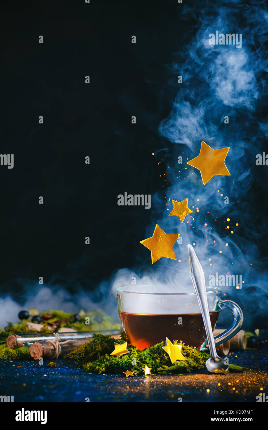 Una tazza di tè con un denso vapore, un antico cucchiaio, stelle dorate e magico polvere pixie su uno sfondo scuro. fiaba concetto con glitter. Foto Stock