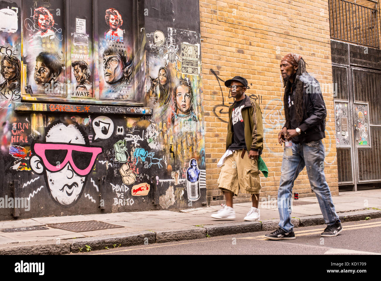 Due uomini afro neri che camminano oltre un muro coperto di graffiti e murales che rappresentano volti di personaggi famosi del cinema a Brick Lane, Shoreditch. Foto Stock