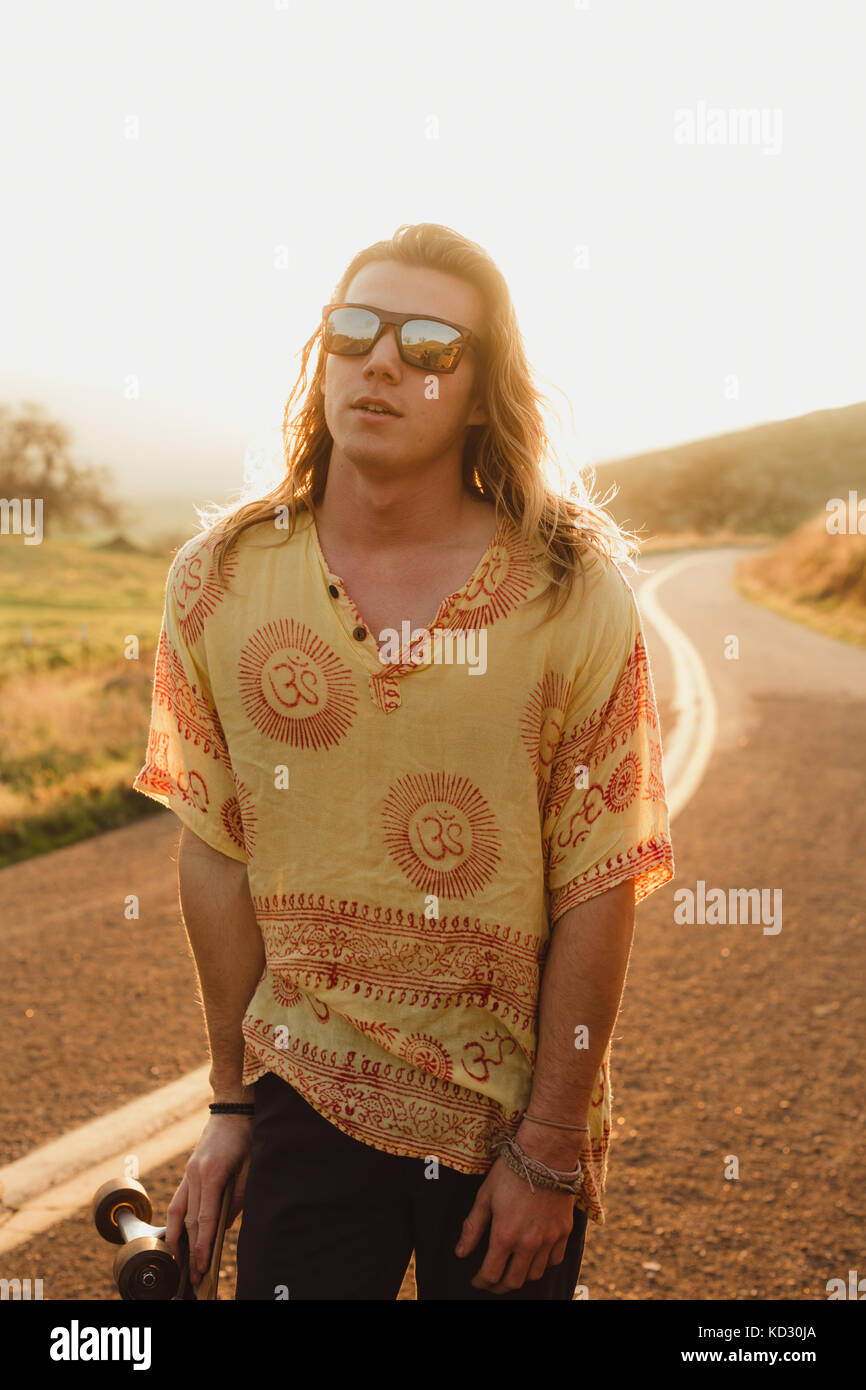 Ritratto di capelli lunghi maschio giovane guidatore di skateboard su strada rurale, Exeter, CALIFORNIA, STATI UNITI D'AMERICA Foto Stock