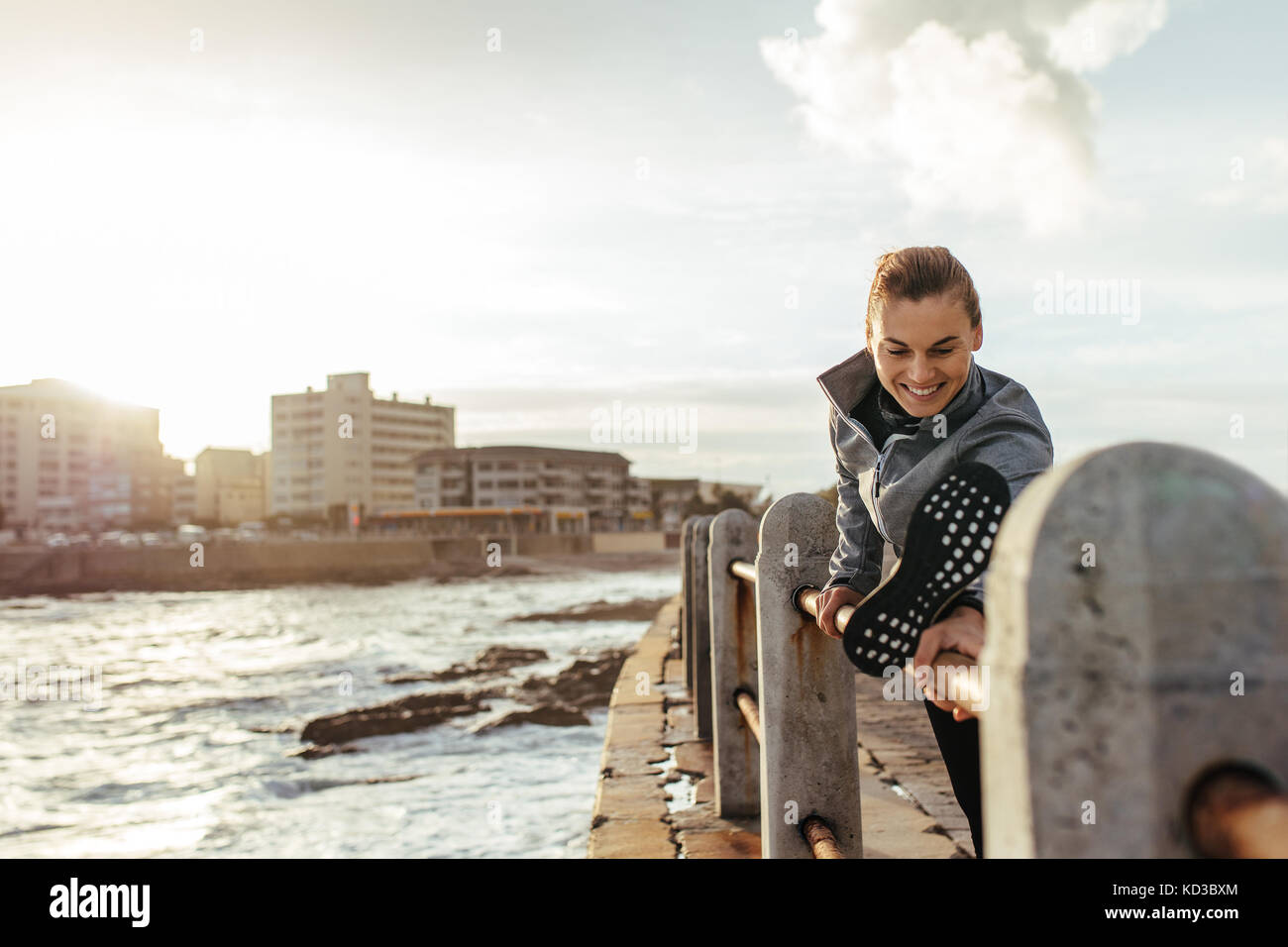Giovane donna sorridente che fa esercizio di stretching delle gambe su ringhiere sulla strada sul mare. fitness donna che avverte sulla strada in riva al mare. Foto Stock