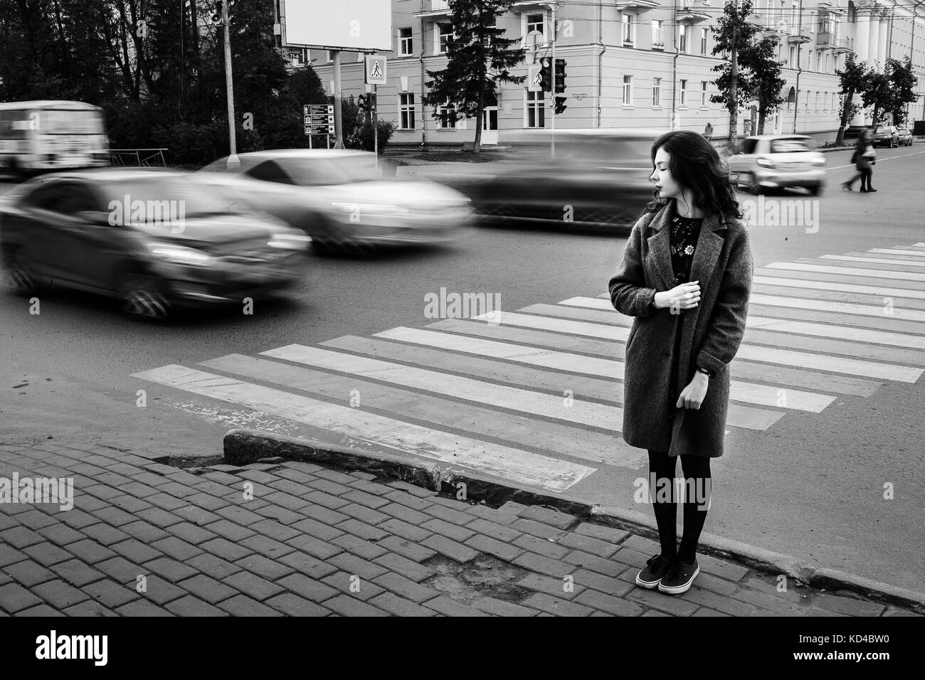 Una ragazza adolescente blue coat permanente al semaforo sulla strada di città su un cloudly giornata autunnale con veicoli che passano da in motion blur. Foto Stock