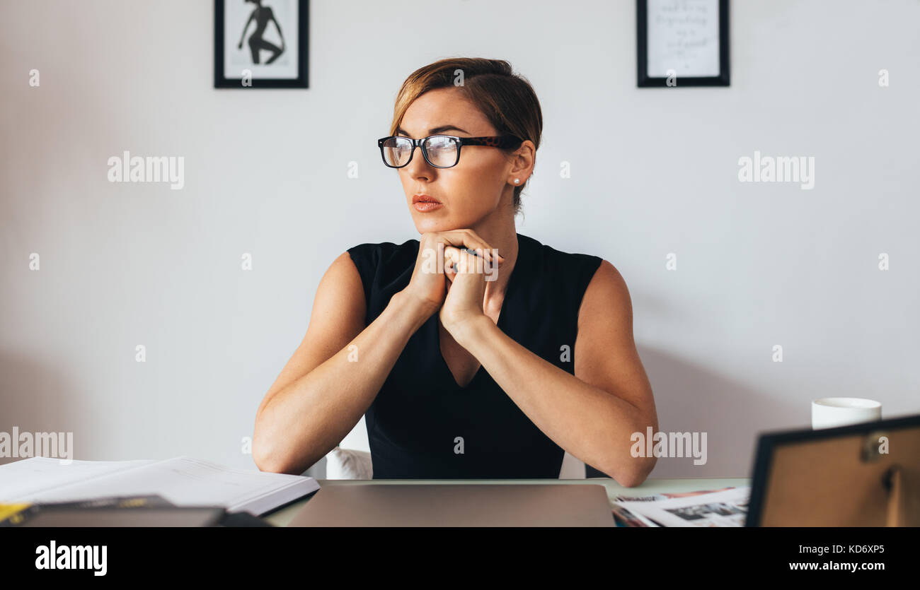 Donna che indossa gli occhiali seduti alla scrivania in ufficio e che guarda lontano. donna d'affari che pensa profondamente con il mento appoggiato sulle mani. Foto Stock