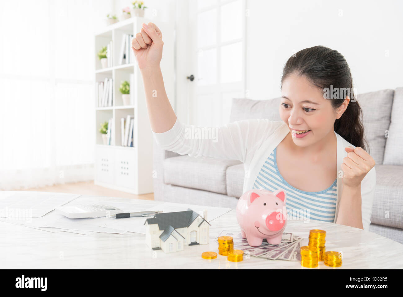 Sorridenti donna risparmio abbastanza fondo può acquistare un nuovo appartamento e seduto in salotto guardando al modello di casa compiendo il gesto di successo celebrazione. Foto Stock