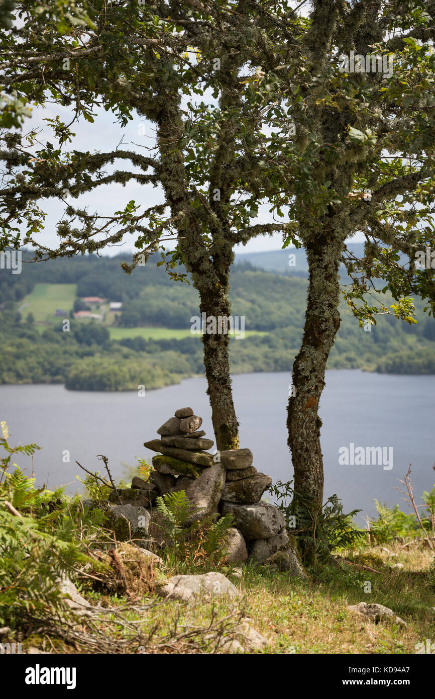 Lac DE VASSIVIERE, FRANCIA - 27 LUGLIO 2017: antica cairns o stacking di roccia sotto un albero di quercia coperto muschio, con il lago sullo sfondo. Foto Stock