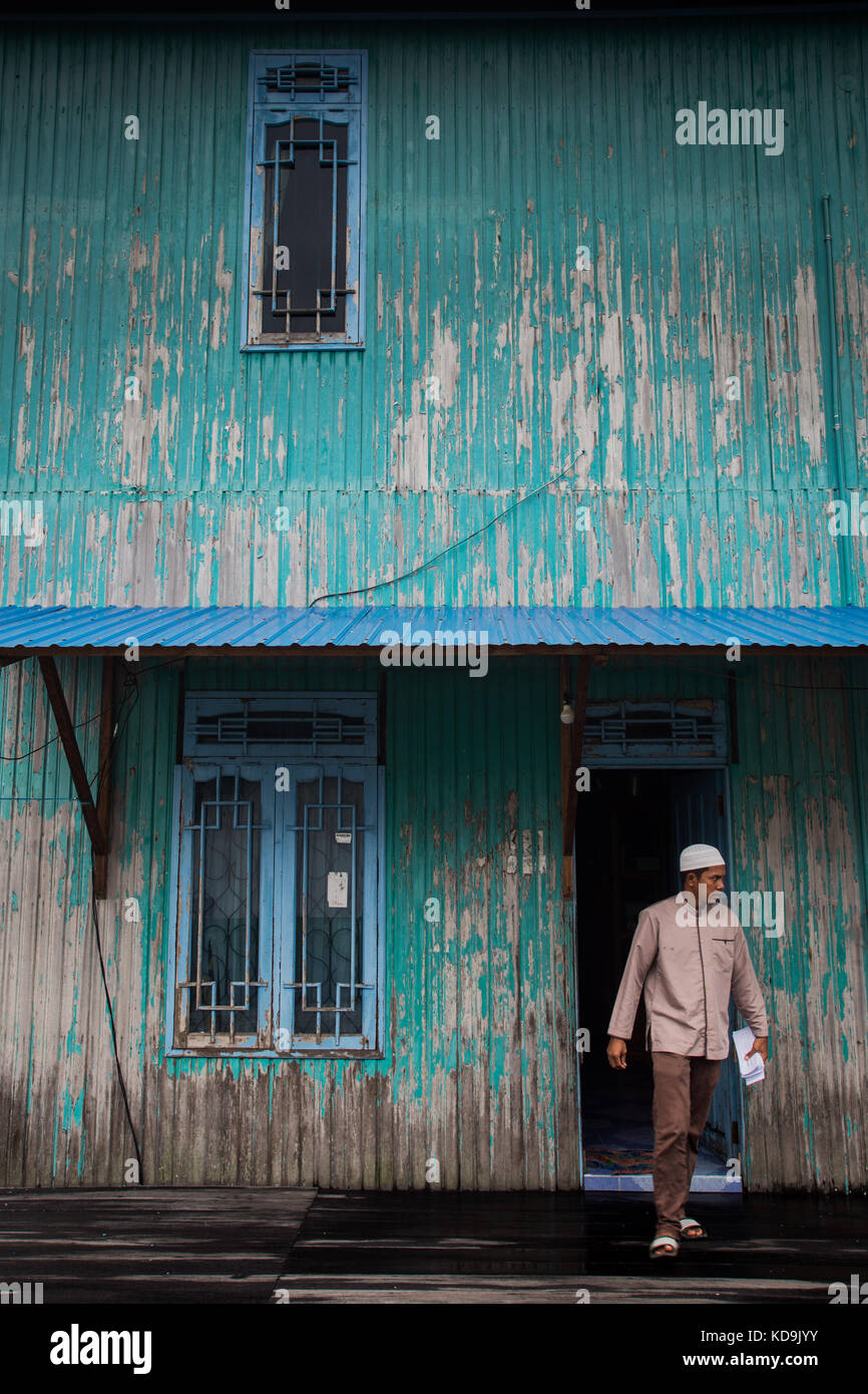 Uomo musulmano lasciando un rustico verniciato blu casa nel Kalimantan, Borneo indonesiano scene di strada Foto Stock