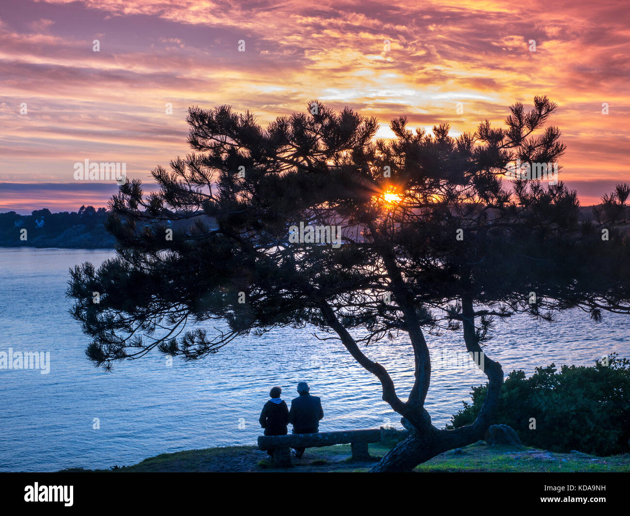 KERFANY LES PINS Coppia matura seduta su panchina in silhouette guardando un colorato tramonto attraverso la baia a Moëlan sur Mer Bretagna Francia Foto Stock