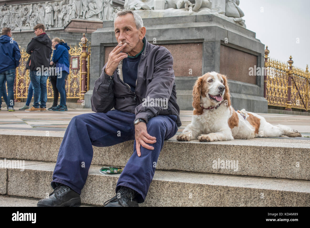 Vecchio / anziani uomo seduto sui gradini del Albert Memorial, fumare una sigaretta, il suo cane che giace accanto a lui. I giardini di Kensington, Londra, Inghilterra, Regno Unito. Foto Stock