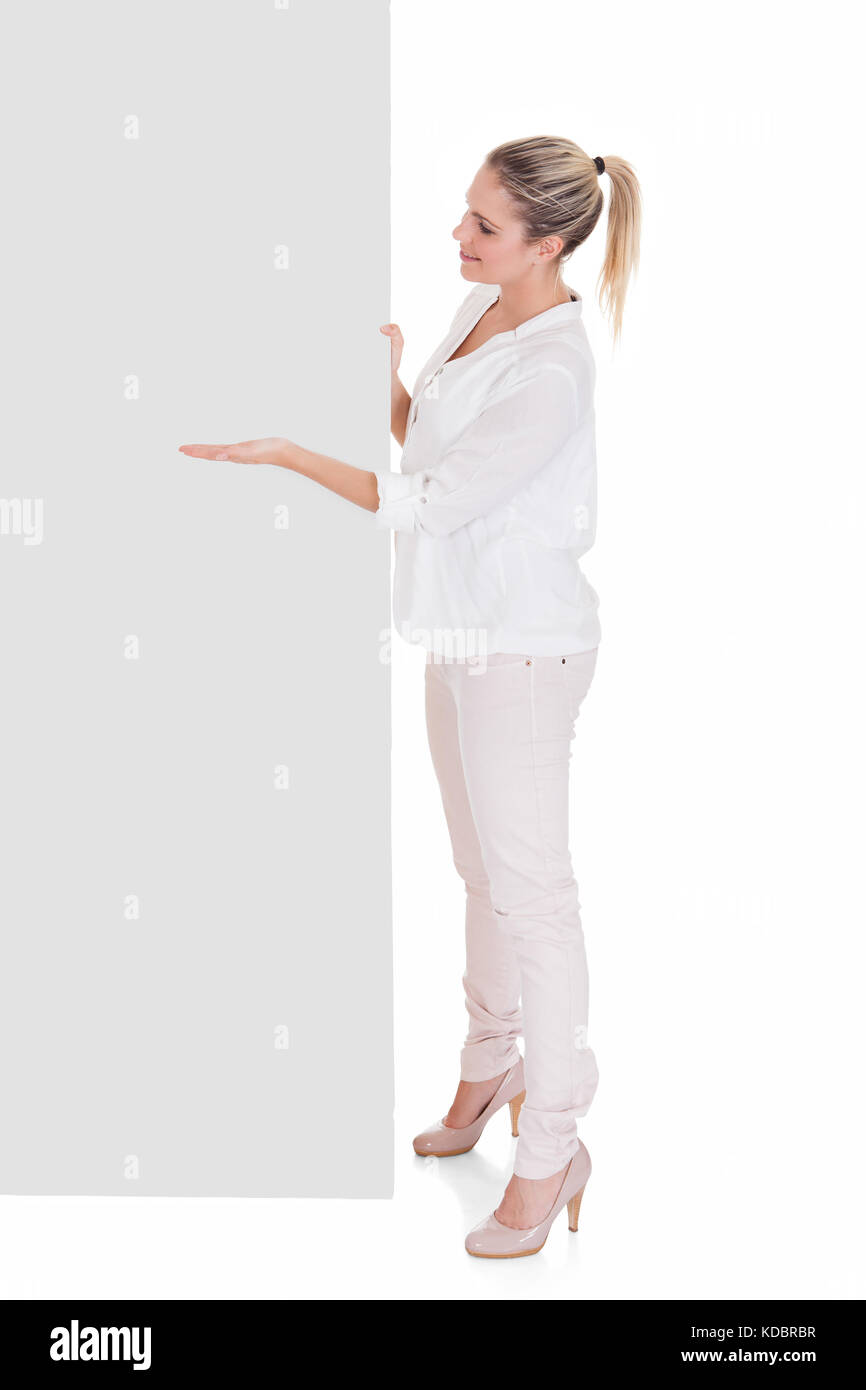 Giovane donna in piedi vicino alla targhetta vuota e presentante su sfondo bianco Foto Stock