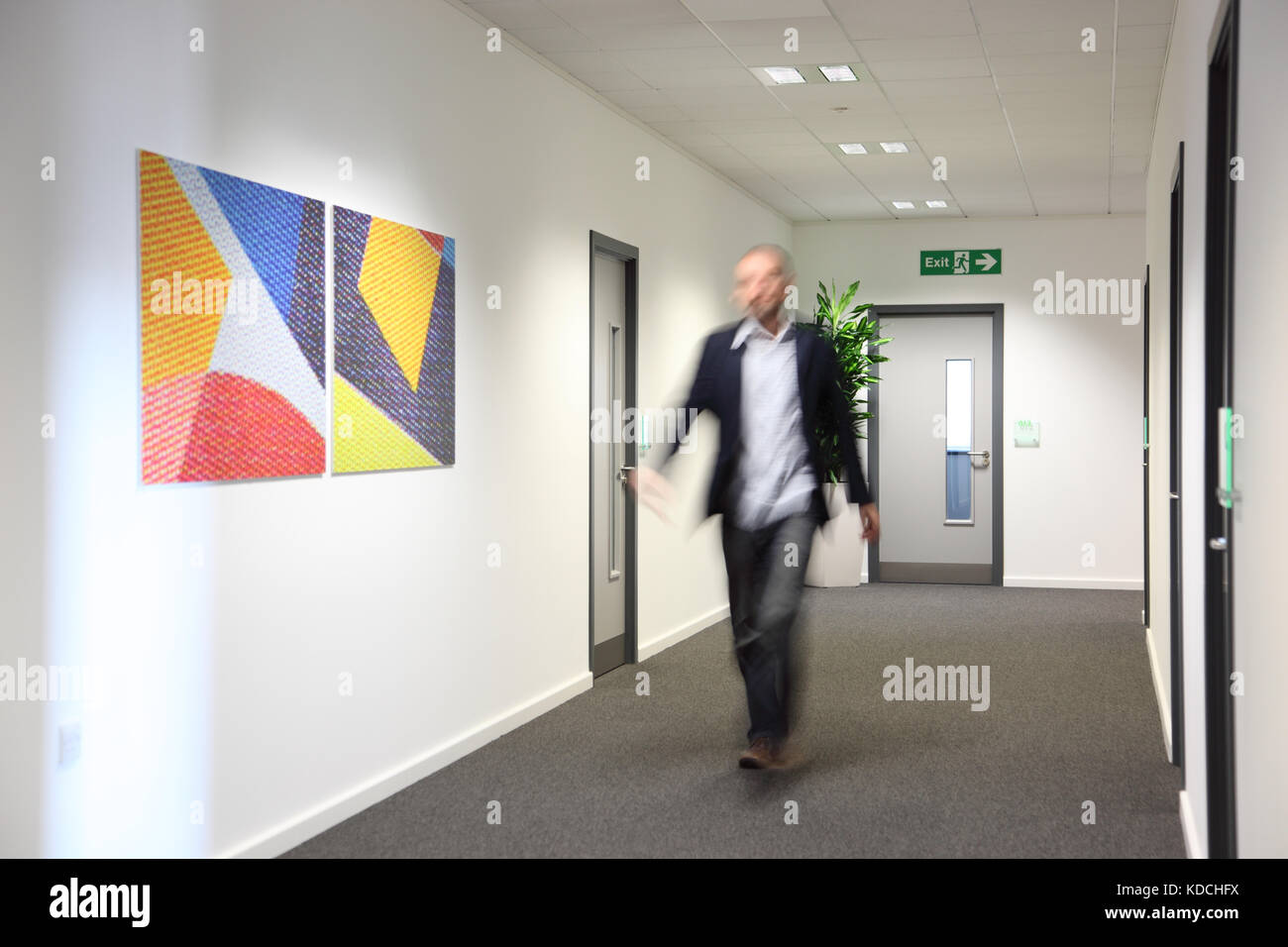 Un vestito in modo informale uomo (sfumata dal movimento) cammina lungo un corridoio in un moderno edificio per uffici. Mostra arte sulle pareti e impianto di pentola in angolo. Foto Stock