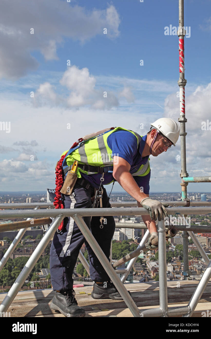 Ponteggiatore erige un lavoro temporaneo di piattaforma su un blocco a torre alto sopra di Londra. Mostra cablaggio di sicurezza nell'uso. Foto Stock