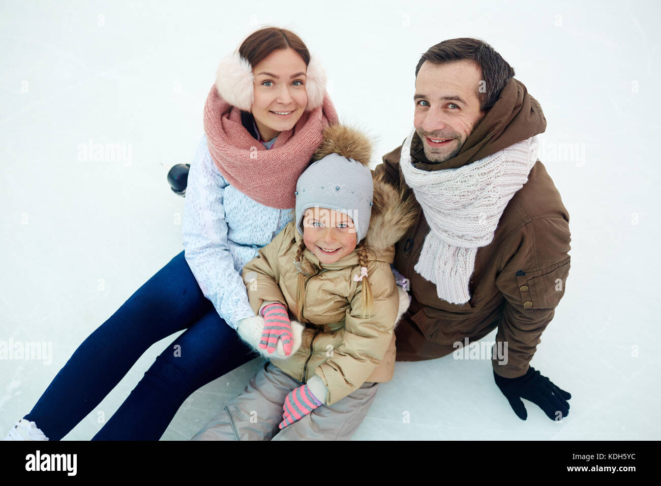 I giovani genitori e la loro figlia in winterwear guardando la fotocamera mentre è seduto sulla pista di pattinaggio Foto Stock