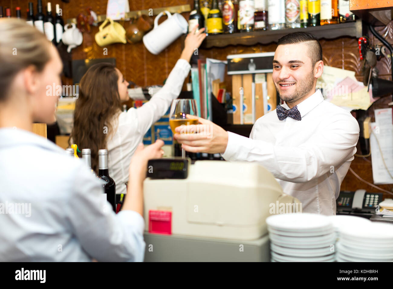 I dipendenti lavorano in un bar: bello barista maschio è dare un bicchiere di vino bianco per una cameriera femmina mentre il barista è di prendere una bottiglia di spiri Foto Stock