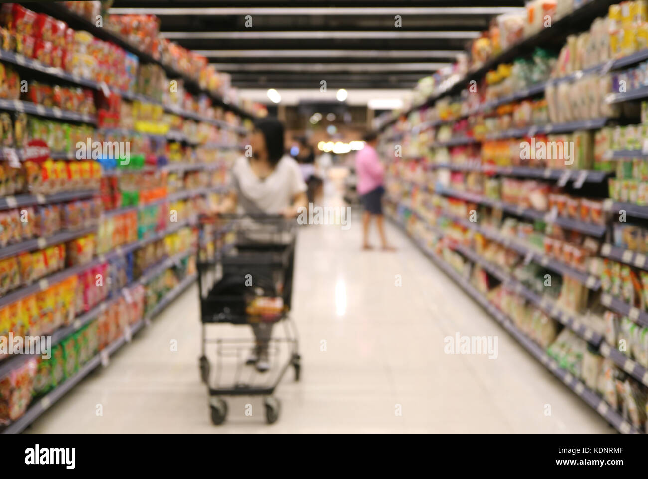 Fuori fuoco colpo di una donna con il suo carrello guardando il ripiano di generi alimentari nel supermercato Foto Stock