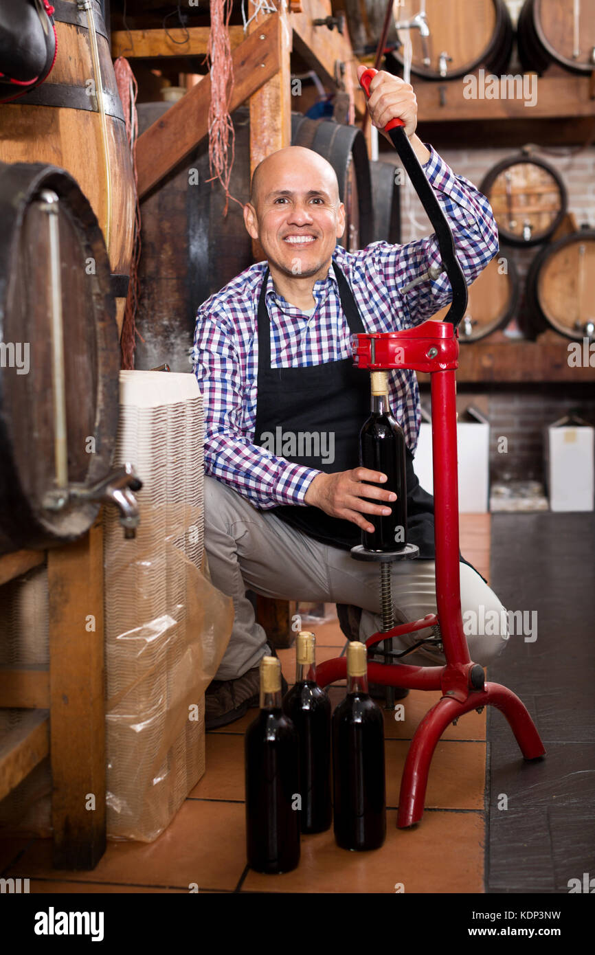 Allegro uomo che indossa il grembiule bottiglia utilizzando apparecchiatura di tappatura in casa del vino Foto Stock