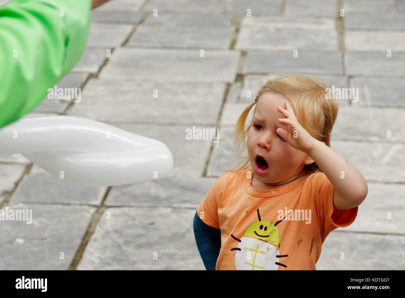 Una bambina di 3 anni) a sbadigliare mentre lei guarda un palloncino scultore rendendo un palloncino. Foto Stock