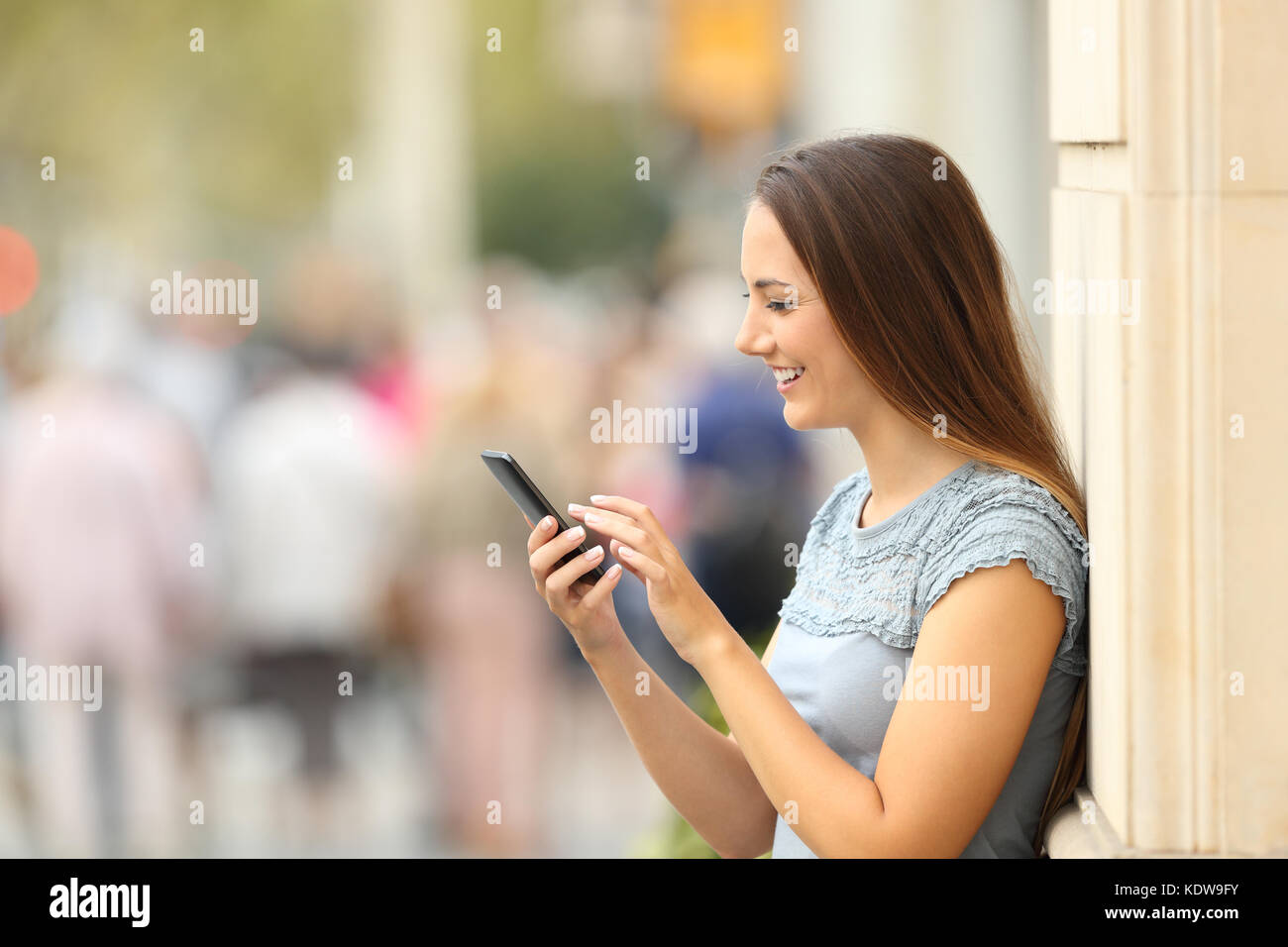 Vista laterale di una ragazza felice controllando il contenuto su uno smart phone appoggiato su di una parete sulla strada Foto Stock