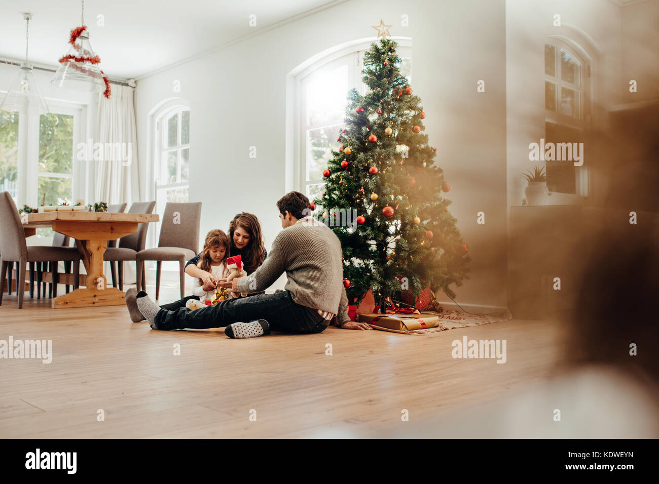 Famiglia seduta vicino ad albero di Natale apertura di confezioni regalo. Coppia giovane aiutando la loro figlia aprire i regali di Natale. Foto Stock