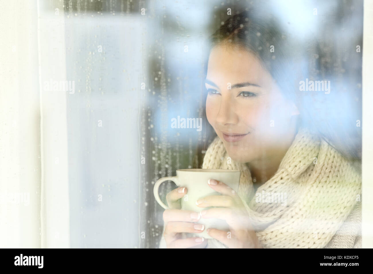 Ritratto di una ragazza di guardare attraverso una finestra tenendo una tazza di caffè in un giorno di pioggia di inverno Foto Stock