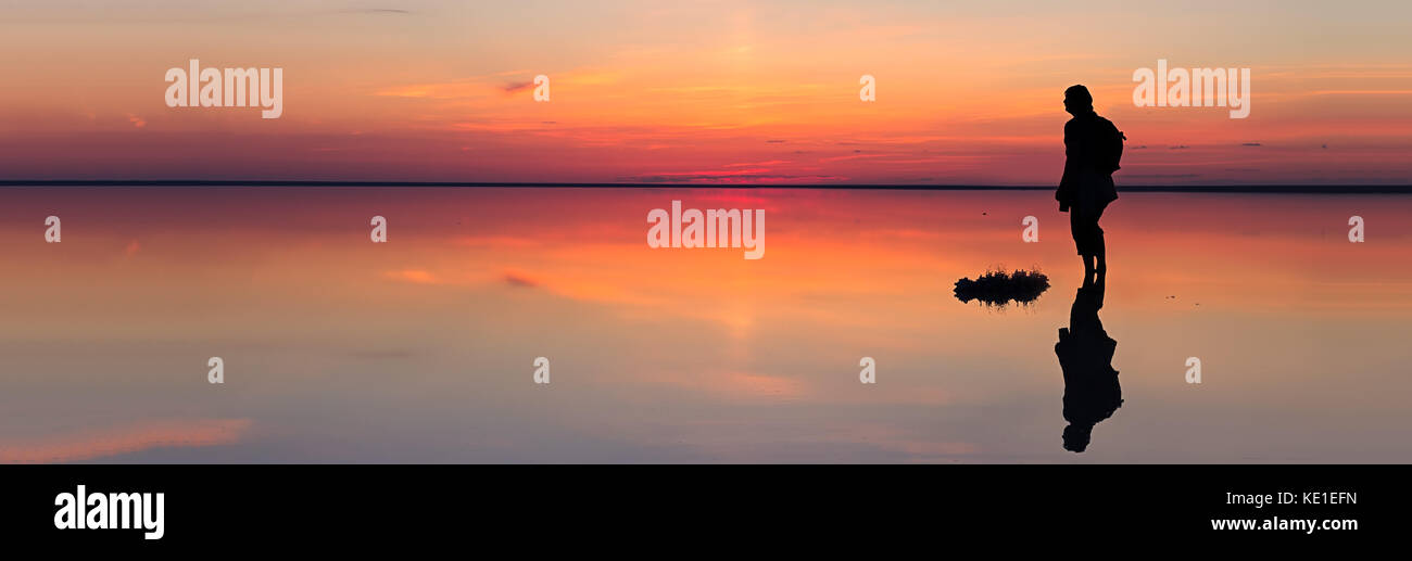 Silhouette di un solo uomo che guarda verso la vibrante tramonto riflesso in acque poco profonde di solt lago dimensioni banner Foto Stock