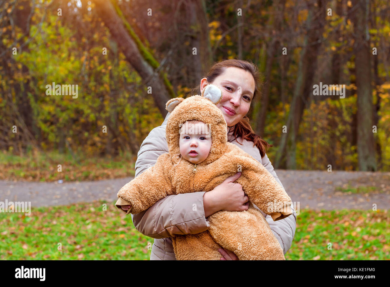Donna con bambino in armi, ponendo sullo sfondo del parco d'autunno. bambino vestito in caldi stilizzata di Teddy bear costume, divertirsi sulla madre di braccia. famiglia w Foto Stock